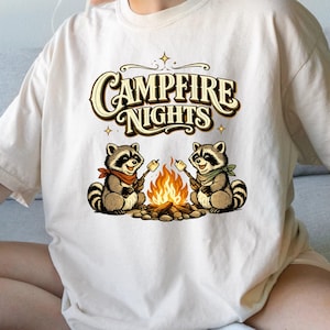 Retro illustration of two raccoons wearing bandanas roasting marshmallows over a campfire with the text Campfire Nights above them on a cream background. — product photo 4