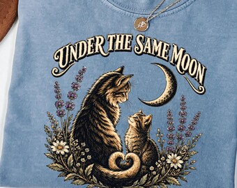 May include: A blue short-sleeve t-shirt featuring a graphic print of two cats sitting among flowers and a crescent moon. The text "UNDER THE SAME MOON" is printed above the cats. The shirt has a crew neckline and a Comfort Colors brand label.
