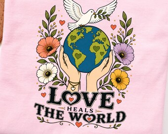 May include: A light pink t-shirt featuring a graphic of two hands holding a globe with a white dove above it. The design includes colorful flowers, small hearts, and the text "LOVE HEALS THE WORLD" in a bold, black, decorative font style.