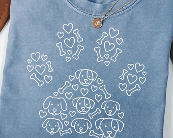 May include: A blue short-sleeve t-shirt featuring a large white graphic of a paw print composed of smaller dog faces, hearts, and bones. The inner neck label reads Comfort Colors, 100% cotton, made in Honduras, size S-CH. A gold necklace rests on the collar.