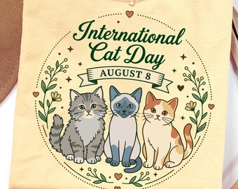 May include: A pale yellow t-shirt featuring a circular graphic with the text "International Cat Day" and "AUGUST 8". The design includes illustrations of three cats, a grey tabby, a Siamese, and a calico, surrounded by a floral wreath and small hearts.