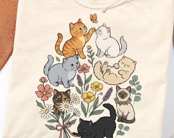May include: A cream-colored t-shirt featuring a graphic print of six kittens in various colors interacting with a bouquet of colorful flowers and a small butterfly. The shirt has a Comfort Colors brand label visible at the inner neckline.