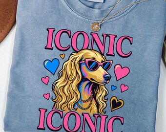 May include: A blue short-sleeve t-shirt featuring a graphic of a long-haired dog wearing pink sunglasses and a gold chain. The text "ICONIC" appears in pink above and below the dog, surrounded by small blue, pink, and gold heart illustrations.