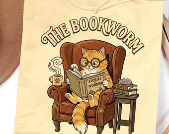 May include: A pale yellow t-shirt featuring a graphic of an orange cat wearing glasses, sitting in a brown armchair while reading a book titled Stories of Wonder. The text The Bookworm is arched above the cat, with a mug and a stack of books nearby.
