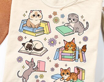 May include: A cream-colored t-shirt featuring a graphic print of six cartoon cats interacting with stacks of colorful books and small flowers. The shirt has a Comfort Colors brand label at the inner neckline and a crew neck collar design.