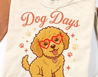 May include: A cream-colored t-shirt featuring a graphic of a golden-brown puppy wearing red heart-shaped glasses and a collar with a heart tag. The text "Dog Days" is printed in a reddish-orange script above the puppy, surrounded by small paw prints.