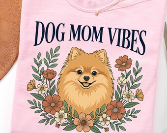 May include: A light pink t-shirt featuring the text DOG MOM VIBES in dark blue lettering above a graphic of a tan Pomeranian dog surrounded by a wreath of brown, orange, and white flowers with green leaves. The shirt has a Comfort Colors label.