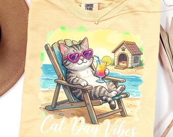 May include: A light yellow t-shirt featuring a graphic of a gray tabby cat wearing pink heart-shaped sunglasses, sitting in a wooden beach chair, holding a tropical drink, with a small doghouse nearby and the text Cat Day Vibes at the bottom.