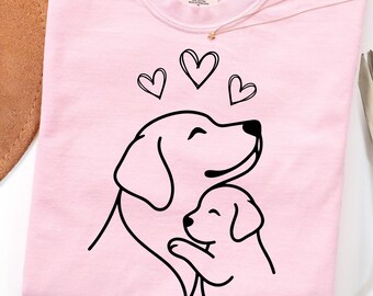 May include: A light pink t-shirt featuring a black line art graphic of an adult dog hugging a puppy with three hearts above them. The shirt has a crew neckline and a Comfort Colors brand label printed on the inside of the collar area.