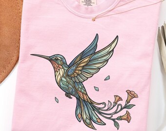 May include: A light pink t-shirt featuring a graphic of a hummingbird with stained-glass style wings in shades of blue, teal, and orange. The bird is surrounded by small floral elements. The inner neck label reads Comfort Colors.
