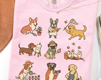 May include: A light pink t-shirt featuring a graphic print of nine different dog breeds engaged in various activities, including gardening, baking, painting, exploring, juggling, and cooking. The shirt has a Comfort Colors brand label at the neck.
