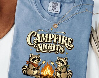 May include: A blue short-sleeve t-shirt with a graphic print featuring two raccoons roasting marshmallows over a campfire. The text "CAMPFIRE NIGHTS" is printed in a stylized font above the raccoons. The shirt has a Comfort Colors brand label.