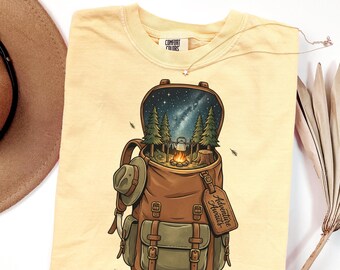 May include: A pale yellow t-shirt featuring a graphic of a brown backpack. The open backpack reveals a forest scene with a campfire, a kettle, and a starry night sky. A small hat hangs on the side, and a luggage tag reads "Adventure Awaits."