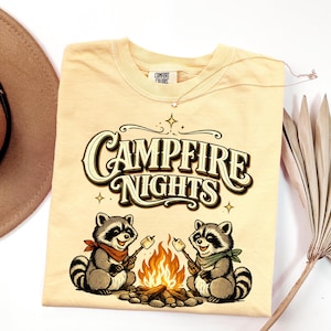Retro illustration of two raccoons wearing bandanas roasting marshmallows over a campfire with the text Campfire Nights above them on a cream background. — product photo 17