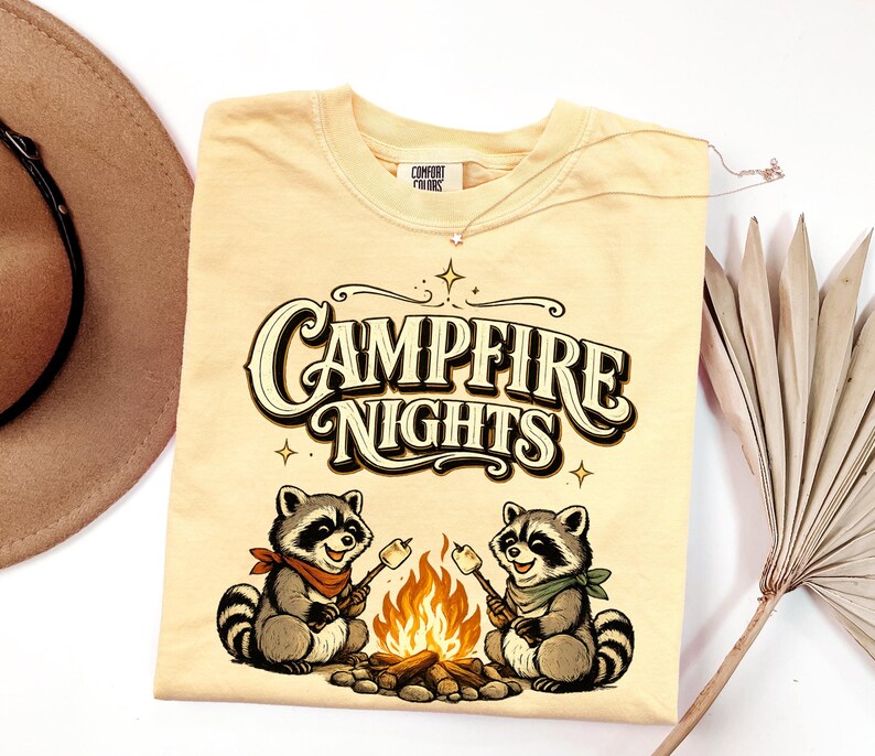 Retro illustration of two raccoons wearing bandanas roasting marshmallows over a campfire with the text Campfire Nights above them on a cream background. — product photo 17