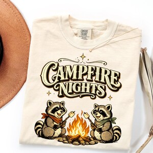 Retro illustration of two raccoons wearing bandanas roasting marshmallows over a campfire with the text Campfire Nights above them on a cream background. — product photo 14