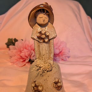 Peut inclure: Une figurine en céramique en forme de cloche représentant une personne portant un chapeau marron à larges bords et une robe blanc cassé mouchetée. La figurine présente un motif floral marron sur le buste et deux sur la base, tenant des fleurs séchées.