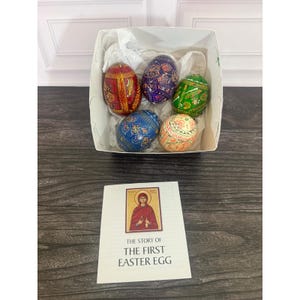 May include: Five hand-painted wooden eggs in a white box with white tissue paper. The eggs feature red, purple, green, blue, and cream colors with intricate patterns. A white card is included with an icon of Mary and the text THE STORY OF THE FIRST EASTER EGG.