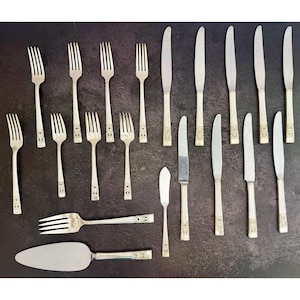 May include: A set of silver-toned flatware including eight forks, seven dinner knives, one butter knife, and one cake server. Each piece features a slender handle with a decorative embossed pattern at the base and a polished metallic finish.