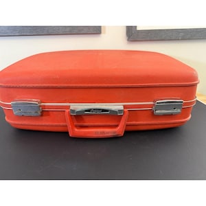May include: A bright orange hardshell suitcase with a textured exterior. The case features a central handle, two silver-toned metal latches, and a silver-toned metal plate on the handle with the text Oshkosh printed in black.