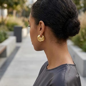 May include: A gold hoop earring featuring a prominent spiral or snail shell design on the surface. The earring has a polished metallic finish and a thick, rounded structure that sits securely on the earlobe, showcasing a detailed circular pattern.