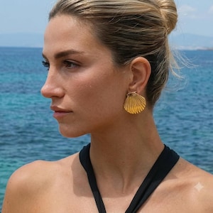 May include: A gold-toned earring shaped like a fan-style seashell with vertical ridges. The earring features a polished metallic finish and a secure backing, designed to sit flush against the earlobe, showcasing a distinct marine-inspired aesthetic.