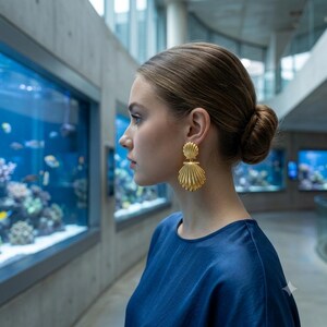 May include: A pair of gold-toned earrings featuring a double-layered shell design. The earrings consist of a smaller, rounded shell top piece connected to a larger, fan-shaped shell bottom piece, both with vertical ridges and a polished metallic finish.