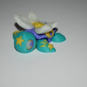 May include: A small, multi-colored figurine featuring a central white flower-like structure with yellow accents. The base is composed of teal rounded components decorated with yellow star patterns and small purple circular details.