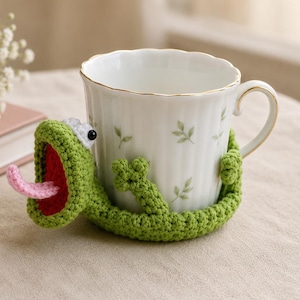 Può includere: Una tazza in ceramica bianca con bordo dorato e motivo a foglie verdi, avvolta da una custodia all'uncinetto a forma di rana verde. La rana presenta una bocca spalancata con interno rosso, una lingua rosa sporgente, occhi neri e braccia.