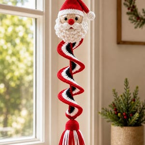 May include: A crocheted Santa Claus ornament featuring a red and white hat, a textured white beard, and a red nose. Below the head is a long, spiraling tail in red, white, and black stripes, ending in a thick red and white yarn tassel for hanging.