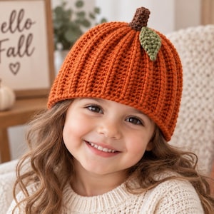 Puede incluir: Un gorro de ganchillo de color naranja brillante diseñado para parecer una calabaza, con textura acanalada vertical, un pequeño tallo marrón en la parte superior y una hoja verde individual unida al lado. El gorro está hecho de hilo tejido.