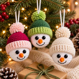 May include: Three crocheted snowman ornaments with white faces, black bead eyes, and orange carrot noses. Each snowman wears a different colored beanie with a matching pom-pom: one in magenta and light pink, one in dark green, and one in cream.