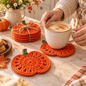 Può includere: Un set di sottobicchieri all'uncinetto a forma di zucca arancione realizzati in filato, con un piccolo stelo verde sulla parte superiore. Un sottobicchiere è posto sotto una tazza in ceramica, mentre una pila di altri quattro è legata con spago.