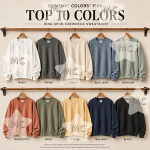 May include: Ten Comfort Colors 1566 ring spun crewneck sweatshirts hanging on two wooden racks. Colors include white, pepper, ivory, blue jean, seafoam, terracotta, moss, butter, true navy, and black, each labeled with its respective color name.