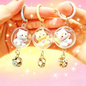 May include: Three keychains featuring clear spherical charms, each containing a small cat figurine in light blue, yellow, or white. Each charm hangs from a coloured plastic ring and includes a gold-toned metal bell with floral cutouts at the base.
