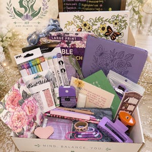 May include: A gift box labeled Wellbeing Mystery Scoops containing a variety of items including a floral notebook, a purple butterfly-embossed case, colorful markers, pens, a hand sanitizer, a hair clip, paper clips, and inspirational cards.