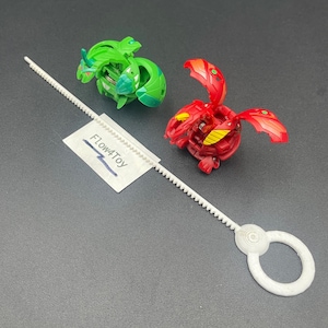 Peut inclure: Deux petites figurines de créatures sphériques transformables en vert et rouge, accompagnées d'une tirette en plastique blanc avec une poignée circulaire. Une petite étiquette en papier blanc porte le texte Flow4Toy imprimé en noir.