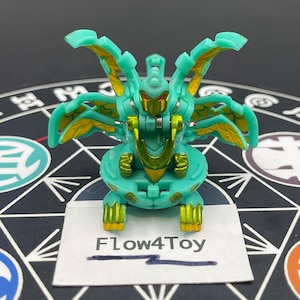 May include: A turquoise and metallic gold Bakugan action figure with multiple wing-like appendages, red eyes, and a central chest plate. A white rectangular card with the black printed text Flow4Toy is positioned directly in front of the figure.