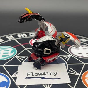 May include: A mechanical dragon-like toy figure in black, red, and silver with a yellow horn and accents. The figure features articulated segments and is positioned behind a white rectangular card with the text Flow4Toy printed in black ink.