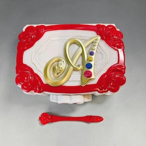 Peut inclure: Un boîtier compact rectangulaire en plastique blanc et rouge avec une grande lettre A dorée sur le couvercle, ornée de quatre petites pierres précieuses colorées incrustées. Un outil en plastique rouge assorti est placé sous le boîtier.