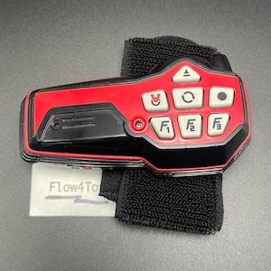 May include: A red and black remote control device with a black fabric wrist strap. The device features six white buttons labeled with symbols and the characters F1, F2, and F3. A small white tag with the text Flow4To is visible underneath the device.