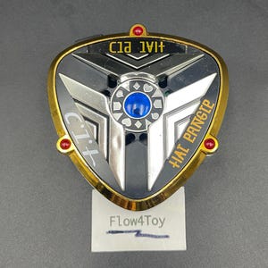 May include: A triangular, metallic gold and black toy device featuring a central blue circular gem, silver geometric accents, and three small red circular lights at the corners. The text "C.I.T." and "HAI PANGTE" is printed on the black surface.