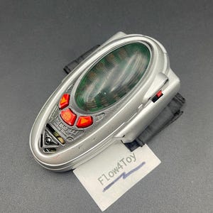 May include: A silver-colored wrist-mounted device featuring a large, oval, dark green translucent lens, three bright red triangular buttons, and a small black strap. The device has a textured gray panel and includes a white tag labeled Flow4Toy.