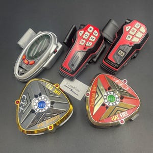 May include: A collection of five plastic toy devices including a silver oval communicator, two red and black rectangular controllers with buttons, and two triangular metallic-finished gadgets labeled C.I.T. and HAT PANGLE with central lights.