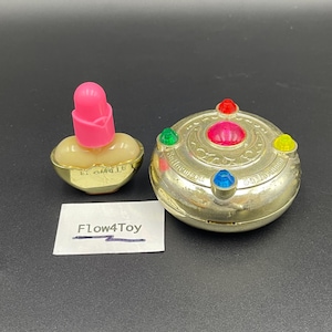 Puede incluir: Un pequeño recipiente dorado en forma de corazón con tapa rosa y un estuche compacto dorado redondo con el texto Sailor Moon, decorado con gemas rojas, verdes, amarillas y azules. Una tarjeta rectangular blanca con el texto impreso Flow4Toy está cerca.