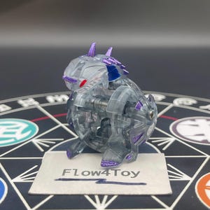 Puede incluir: Una figura de criatura de plástico gris translúcido y morado con ojos rojos y púas en la espalda. La figura está colocada sobre una tarjeta rectangular blanca con el texto Flow4Toy impreso en tinta negra, sobre un diseño geométrico.