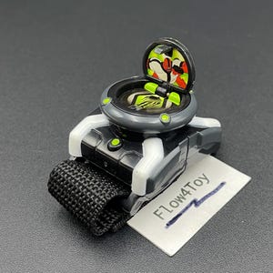May include: A black and grey toy watch with a white strap attachment and a hinged circular lid that opens to reveal a graphic of a red and white character. The device features a black woven fabric band and a small white tag labelled F1ow4Toy.