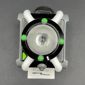 May include: A wrist-worn toy featuring a circular black dial with four bright green circular accents and a central translucent face. The device has white curved side bars, a grey rectangular base, and a small white tag labelled FlowToy.