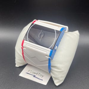 Puede incluir: Un reloj digital blanco con detalles laterales en azul y rojo, que presenta una pantalla rectangular negra con un logotipo circular central. La correa tiene un diseño geométrico moldeado y se exhibe sobre un cojín blanco con la etiqueta Flow4Toy.