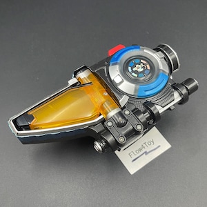 以下が含まれることがあります： 黒、銀、青のプラスチック製おもちゃのデバイス。半透明の琥珀色の三角形のセクション、中央にグラフィックがある円形のダイヤル、小さな赤いボタンが特徴です。Flow4Toyというテキストが書かれた白いタグがベースに取り付けられています。